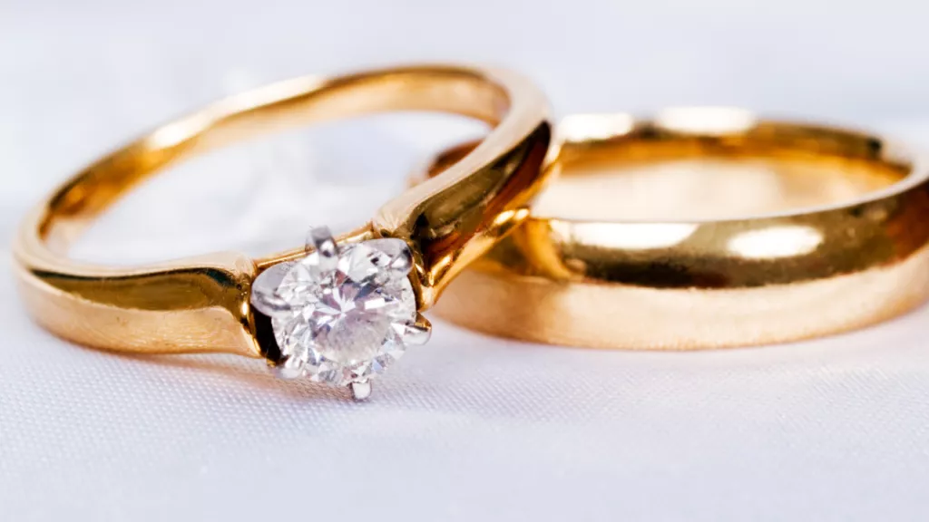 gold engagement ring with a sparkling diamond next to a matching gold wedding band, both resting on a soft white surface.
