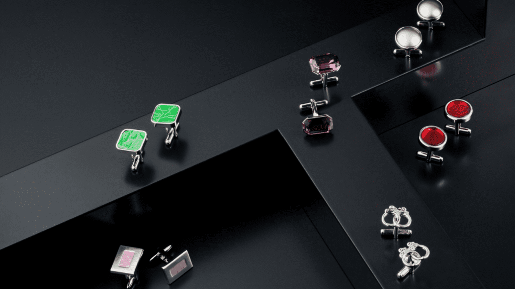 A collection of elegant cufflinks displayed on a sleek black surface, featuring designs in green, purple, red, and silver, arranged in a geometric pattern.