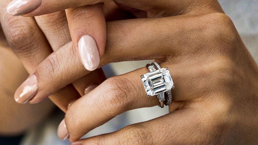 Close-up of a hand wearing a stunning emerald-cut diamond ring with side baguette diamonds, paired with a diamond-studded wedding band.