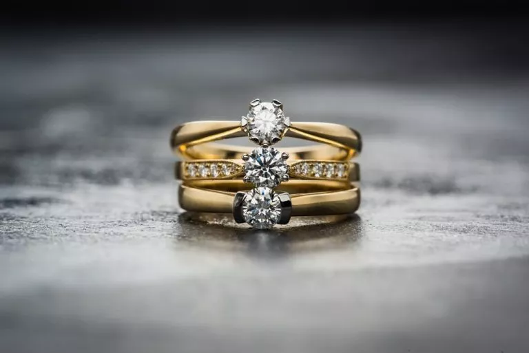 A set of gold engagement rings with brilliant round diamonds, stacked vertically on a textured dark surface, highlighting their richness.