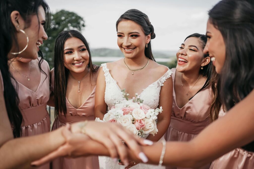 A bride in a white lace gown holds a pink and white rose bouquet, surrounded by bridesmaids in blush dresses, all laughing and sharing a joyful moment.