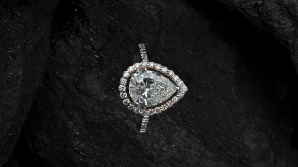 A stunning pear-shaped diamond ring set against a textured, dark background. The central diamond is encircled by a halo of smaller diamonds, which extend onto the band, enhancing its brilliance and elegance. The contrast between the sparkling diamonds and the dark, rugged surface accentuates the ring's exquisite craftsmanship and radiant beauty.