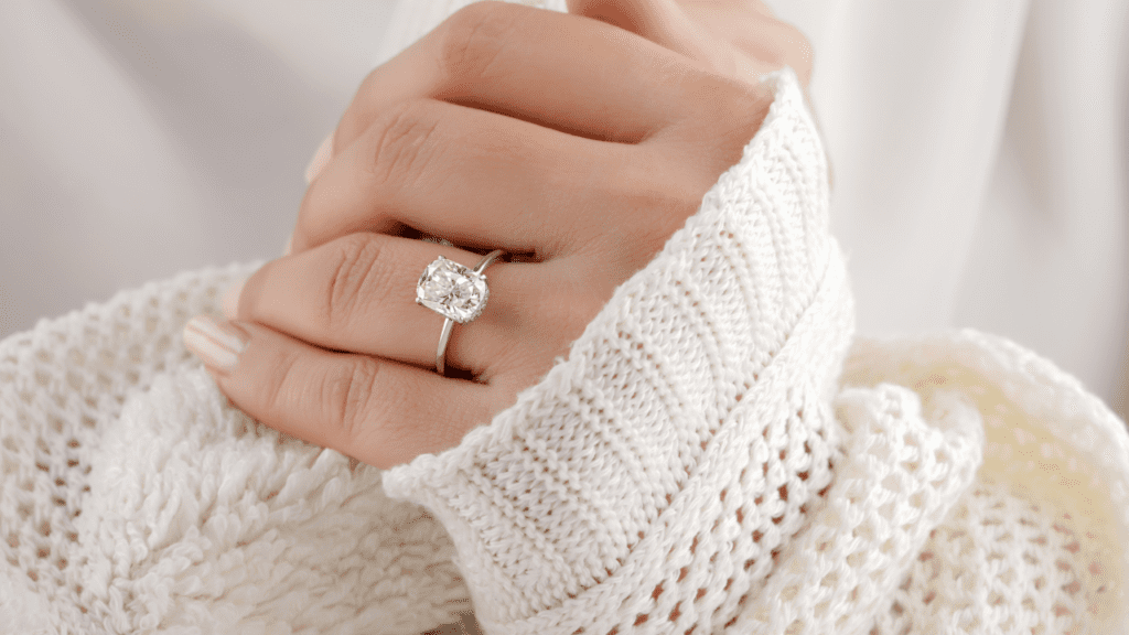A hand wearing a cushion-cut diamond ring, against a cozy white knitted sweater, showcasing its sparkle and adding warmth to the scene.