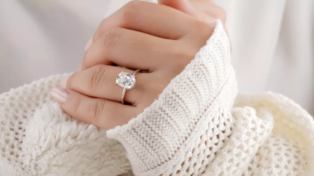 A hand wearing a cushion-cut diamond ring, against a cozy white knitted sweater, showcasing its sparkle and adding warmth to the scene.
