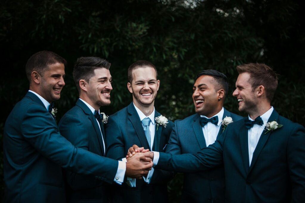 A group of five men dressed in matching blue suits and white shirts are standing together outdoors, sharing a moment of laughter and camaraderie. They are clasping each other's hands in a joyful and celebratory manner. Each man is wearing a boutonniere with white flowers on their left lapel, and three of them are sporting blue bow ties while two have blue neckties. The background is filled with lush greenery, adding to the lively and festive atmosphere of the scene.