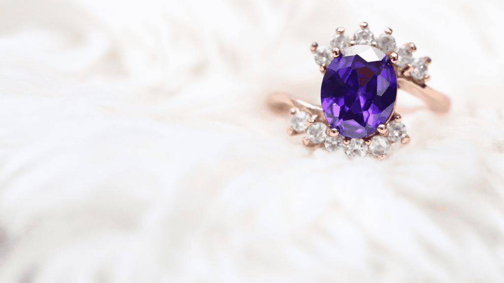 A rose gold ring with an oval purple gemstone, surrounded by sparkling diamonds, rests on a soft, white, fluffy background, exuding elegance.