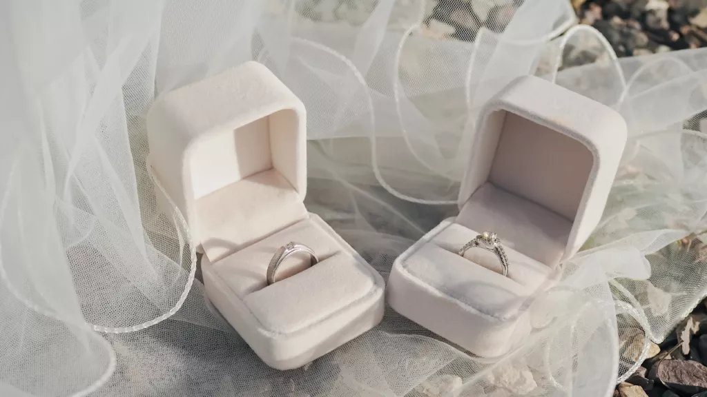 Two velvet boxes hold rings: one with a simple band and small diamond, the other with a detailed ring and central gemstone. Sheer fabric adds a romantic touch.