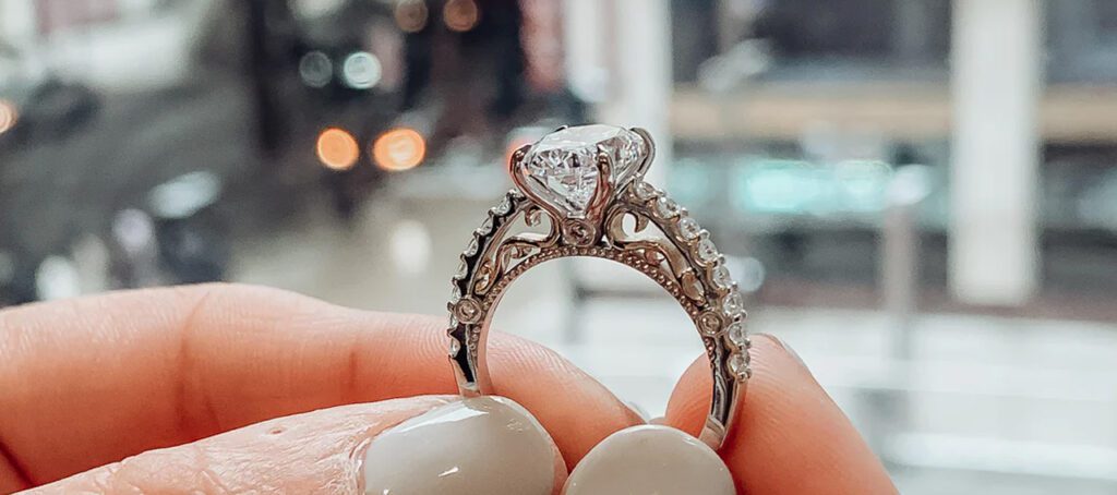 Close-up of a hand holding an ornate engagement ring with a brilliant-cut diamond, against a blurred cityscape background with glowing streetlights.