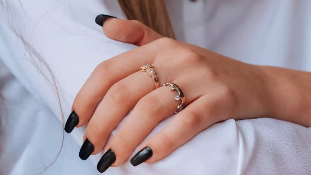 Close-up of a hand with black nail polish, showcasing two elegant rings: a gold ring with intricate design and a silver ring adorned with stones.