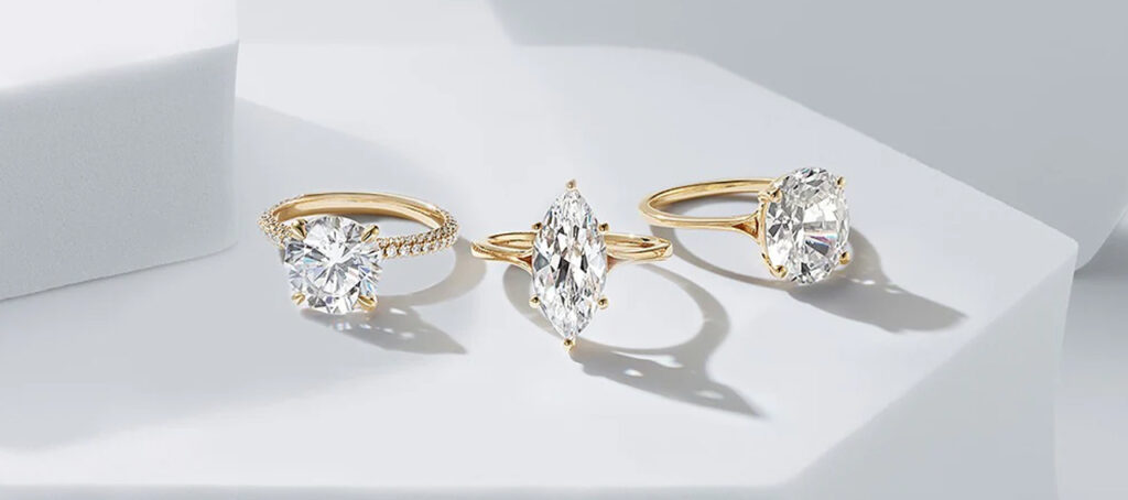 ChatGPTThree gold engagement rings, each with a distinct diamond cut—round, marquise, and oval—are elegantly displayed on a white geometric surface.