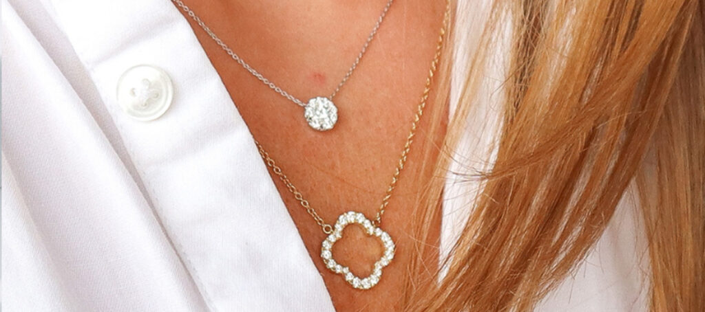 Close-up of a woman's chest wearing a white shirt with two layered necklaces, one with a small round pendant and the other with a larger floral-shaped pendant.