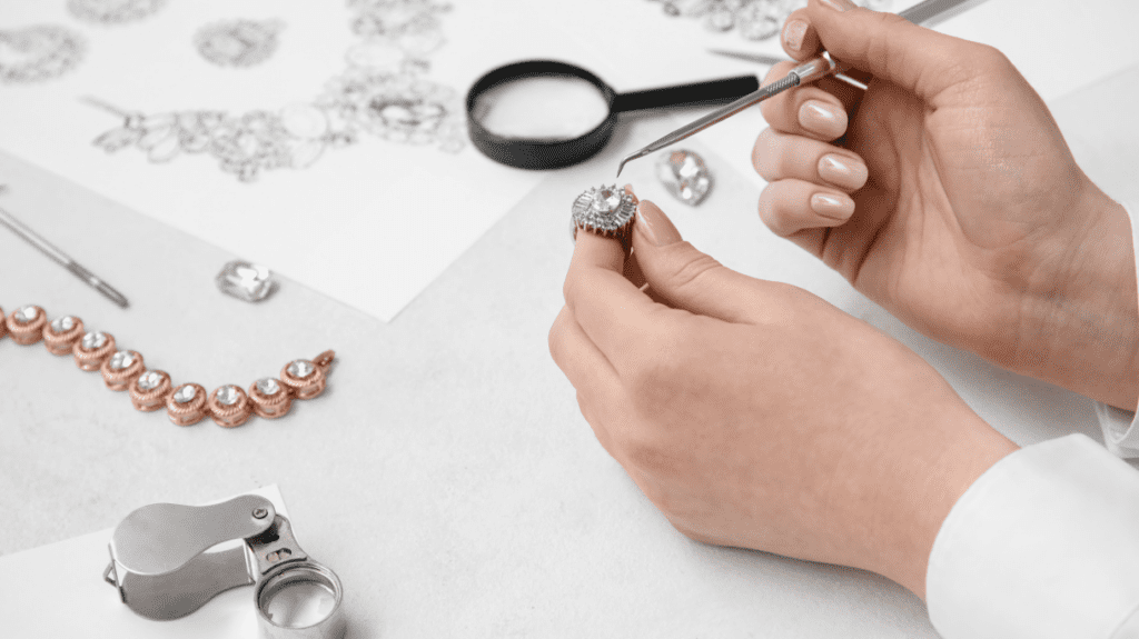 A jeweler carefully examining a diamond ring with precision tools, surrounded by design sketches and a gemstone bracelet on a white workspace.