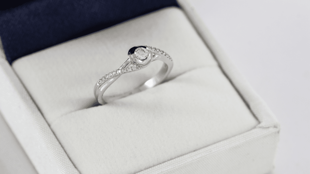 A diamond ring with a round solitaire centerpiece and small diamonds set along the band, displayed in a white velvet ring box.