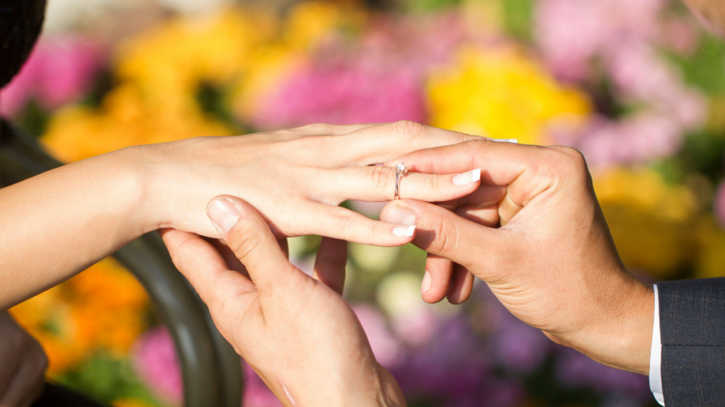 A close-up of a person placing a diamond ring on another person's finger, set against a backdrop of vibrant, blurred flowers in a garden.