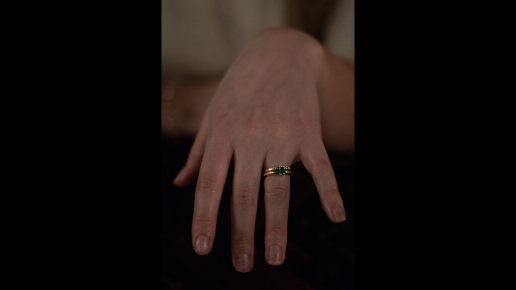 Hand wearing a gold band with a small emerald stone, positioned against a soft-focus background, highlighting the simplicity and elegance of the ring.