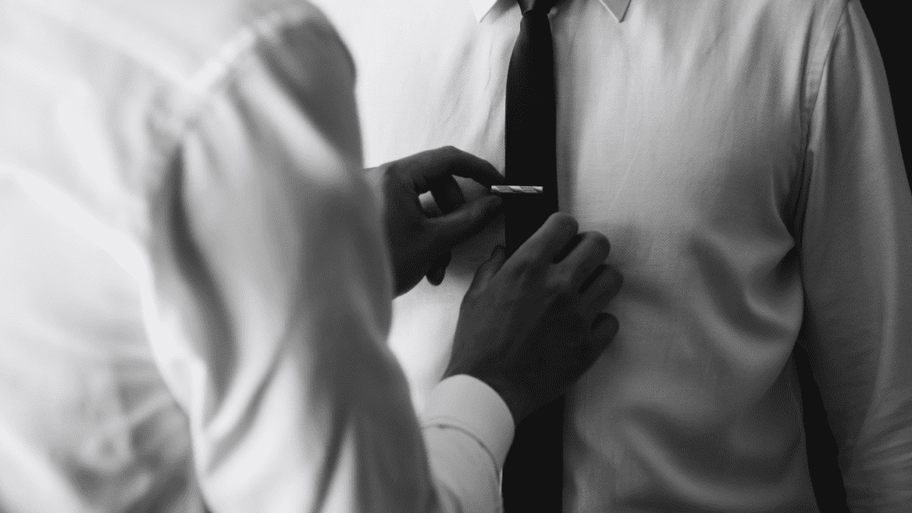 A black-and-white image showing a person adjusting another's tie, emphasizing the elegance and formality of a neatly fastened tie clip on a dark tie.