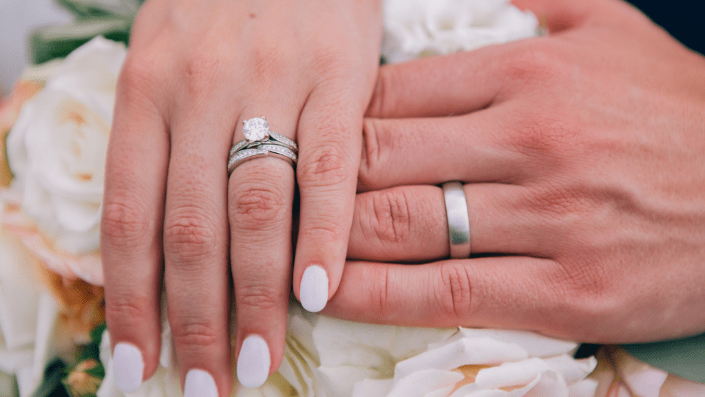 Bride and groom's hands rest on white roses, showcasing her sparkling engagement and wedding rings alongside his simple silver band, symbolizing union.