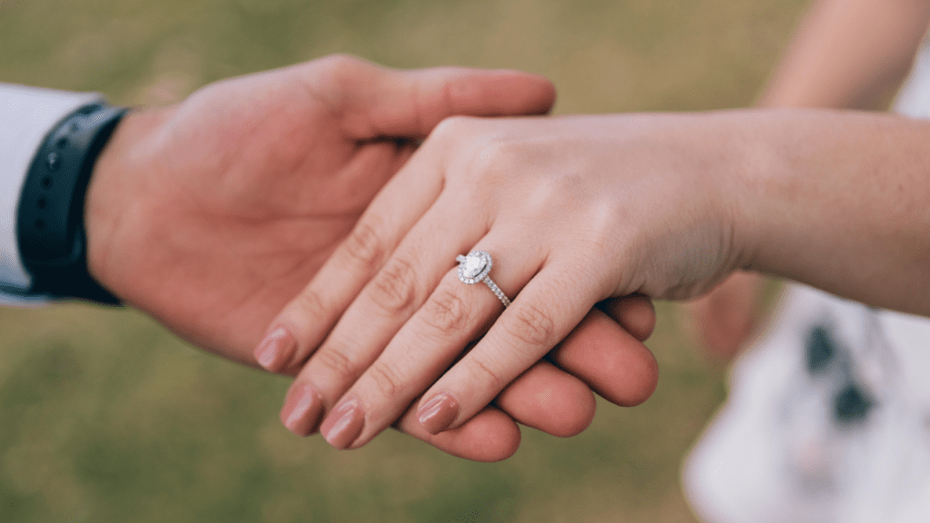 A close-up of a couple holding hands, showcasing a sparkling engagement ring with a halo setting on the woman's hand, highlighting their love and commitment.
