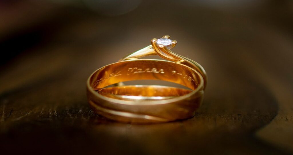 A close-up of intertwined gold rings with an engraved inscription, featuring a single small diamond on top, set against a wooden background.