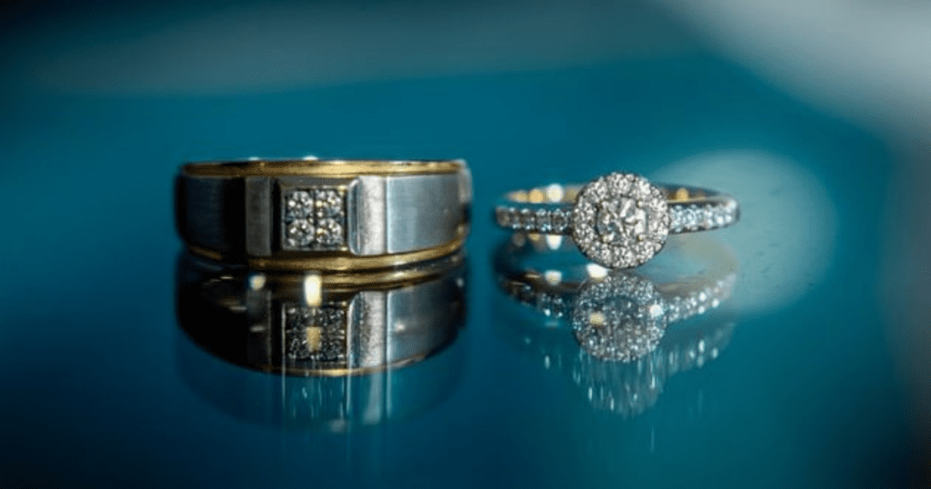 A close-up of two rings on a reflective blue surface. One is a thick gold band with square diamonds, the other a delicate ring with a halo of diamonds.