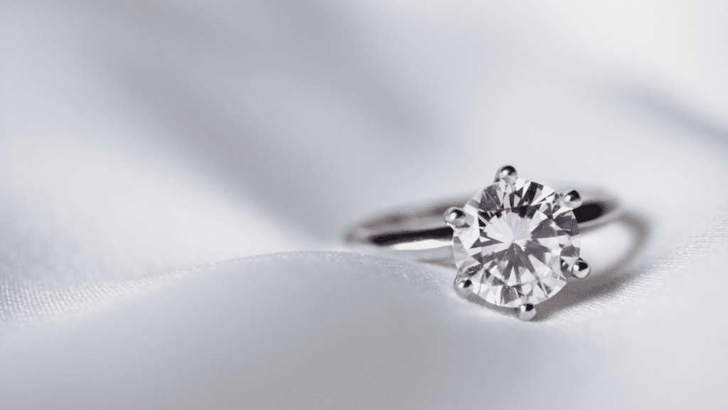 A close-up of a solitaire diamond ring with a round-cut gem set in a simple prong setting, displayed elegantly on a soft white fabric.