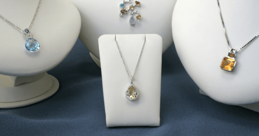 Three elegant necklaces with vibrant gemstones are showcased on white display stands. The center necklace features a pear-shaped green stone.