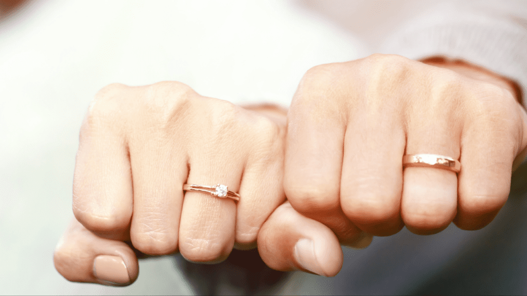 A close-up of two hands showing matching rose gold rings, one with a single diamond solitaire and the other a simple band, symbolizing unity and love.