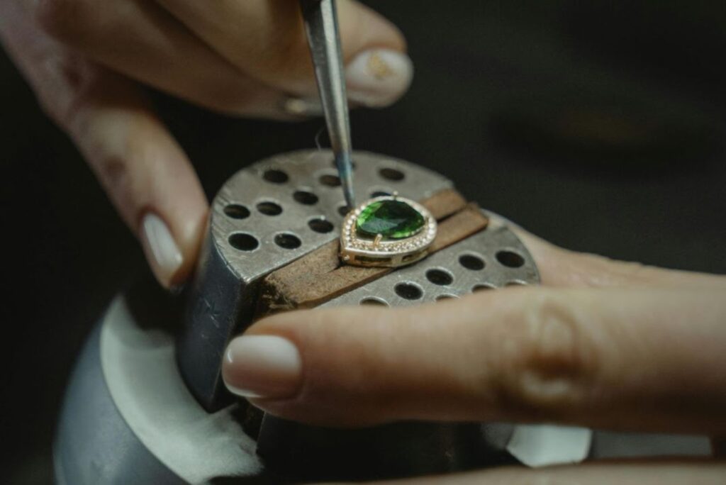 A jeweler meticulously sets a green gemstone into a pendant using precision tools, highlighting the intricate craftsmanship involved in fine jewelry making.