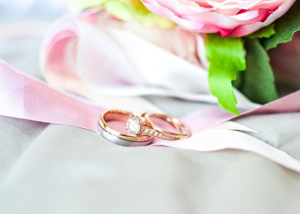 A close-up of two wedding rings on a white background with a pink ribbon and a bouquet of flowers in the background.