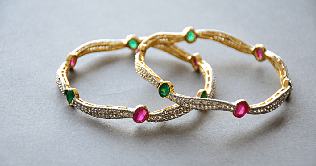 Two ornate gold bangles with sparkling diamond accents, featuring alternating green and pink gemstones, displayed on a sleek gray surface.