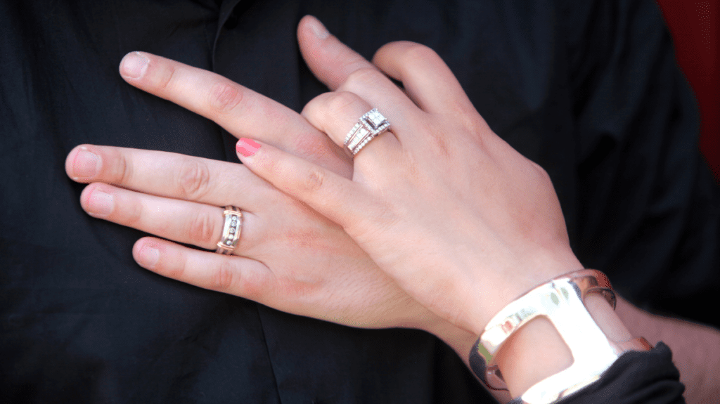 Hands clasped together, showcasing matching wedding bands and a sparkling engagement ring, symbolizing unity and lifelong commitment.