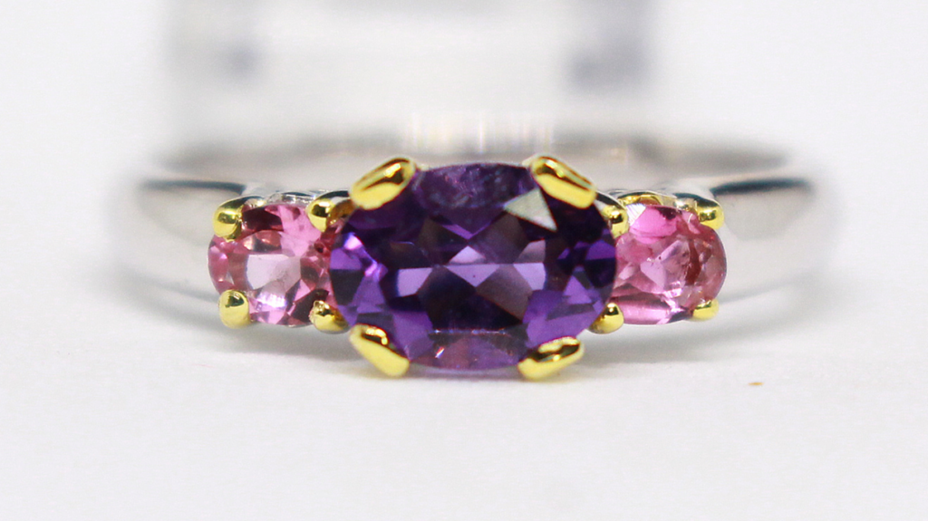 A stunning ring featuring a central purple amethyst flanked by two pink sapphires, set in a silver band with gold prongs, highlighting its vibrant colors.