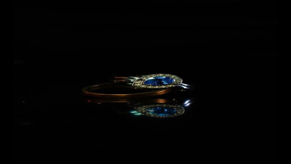 Close-up of a blue gemstone ring with a pear-shaped center stone surrounded by smaller diamonds, set against a dark background with a reflection.