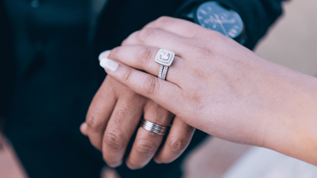 A close-up of a couple holding hands, showcasing their wedding rings. The woman's ring is a square diamond with a double band, the man's a simple band.