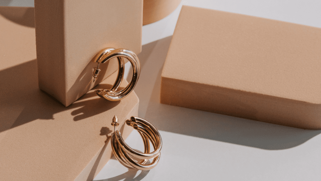 Close-up of a pair of gold hoop earrings resting on beige geometric shapes, casting soft shadows in natural sunlight.