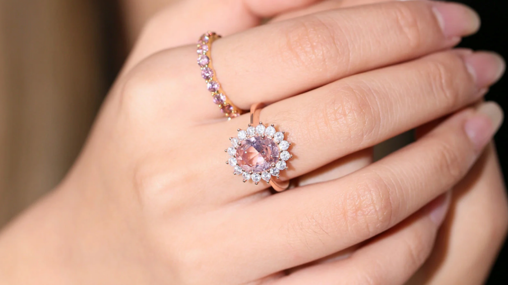 A hand wearing a stunning pink gemstone ring surrounded by a halo of diamonds on one finger and a delicate pink-studded gold band on the other.