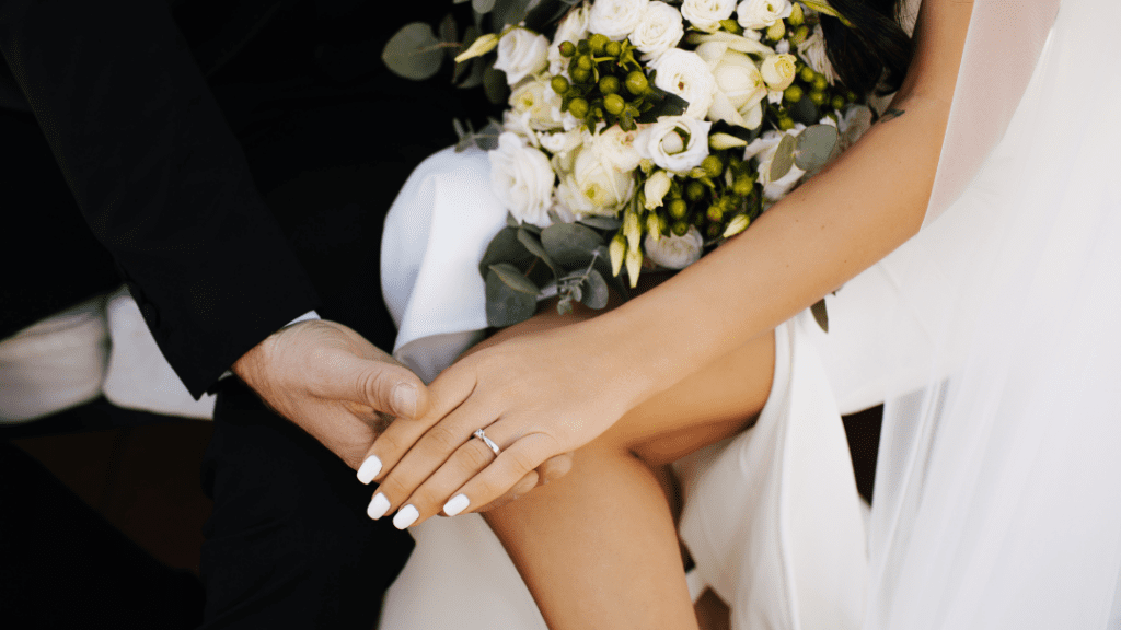 A newlywed couple holds hands, the bride's elegant diamond ring displayed. She holds a bouquet of white roses and green berries, dressed in white.