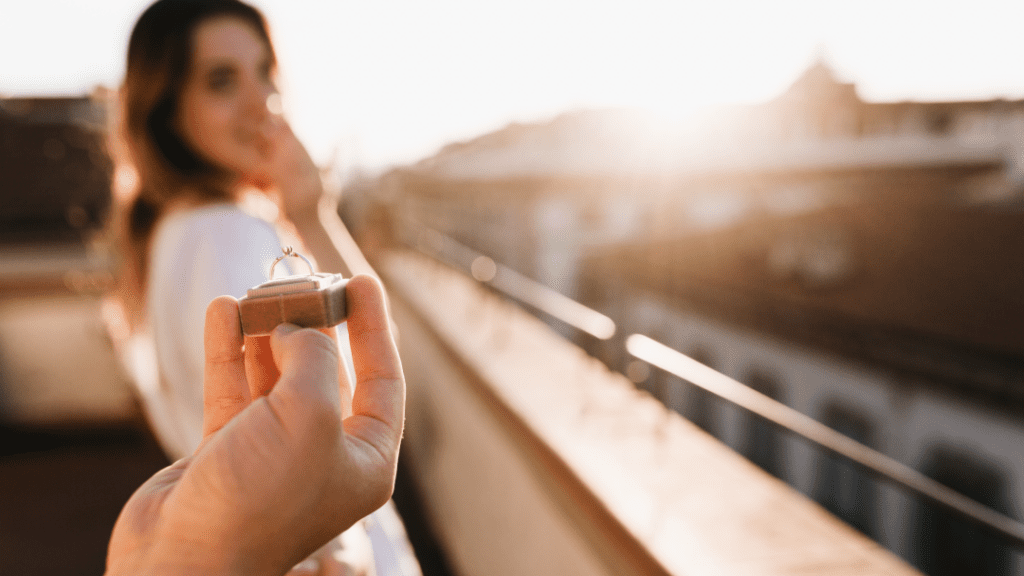 A hand holds out a ring box with a diamond engagement ring, as a blurred woman in the background reacts with joy on a sunny rooftop.