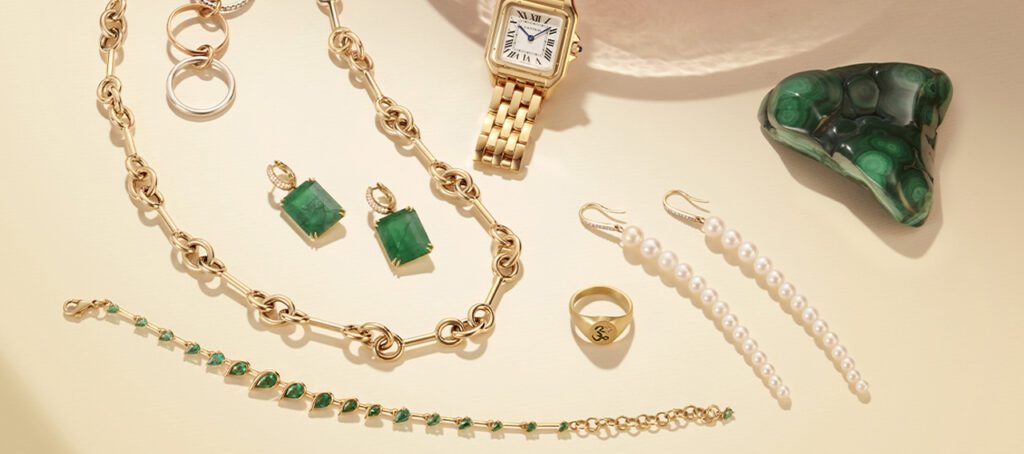 A stylish ensemble of gold jewelry featuring a watch, green gemstone earrings, pearl drop earrings, and a gold bracelet, artfully arranged on a soft surface.