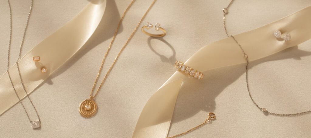 Delicate gold and silver jewelry, including necklaces, rings, and earrings, elegantly displayed with a satin ribbon on a soft beige background.