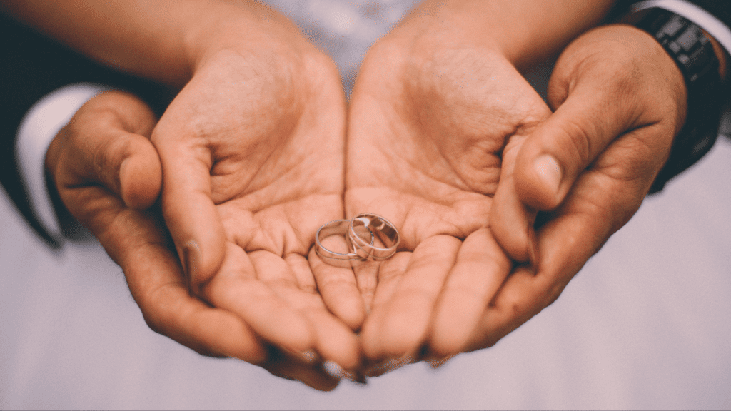 A close-up of two hands, one with a wedding ring on the ring finger, holding two wedding rings.
