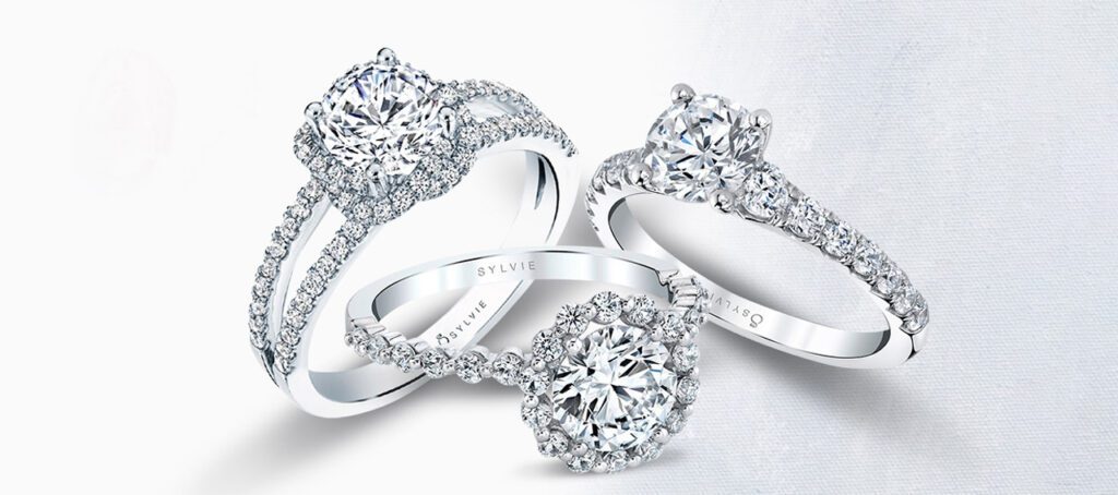 Three dazzling diamond engagement rings, each featuring intricate band designs and brilliant-cut center stones, engraved with "SYLVIE" on the inner band.