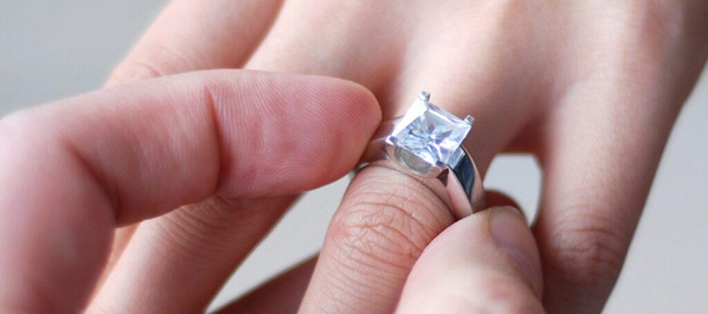 Close-up of a hand placing a silver ring with a square-cut diamond onto another person's finger, capturing a moment of engagement or proposal.