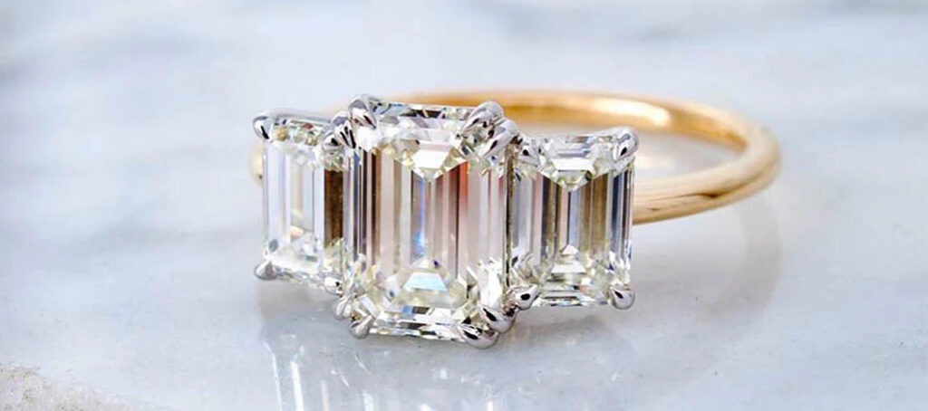 A stunning engagement ring with a trio of emerald-cut diamonds set in a delicate gold band, elegantly displayed on a light marble surface.