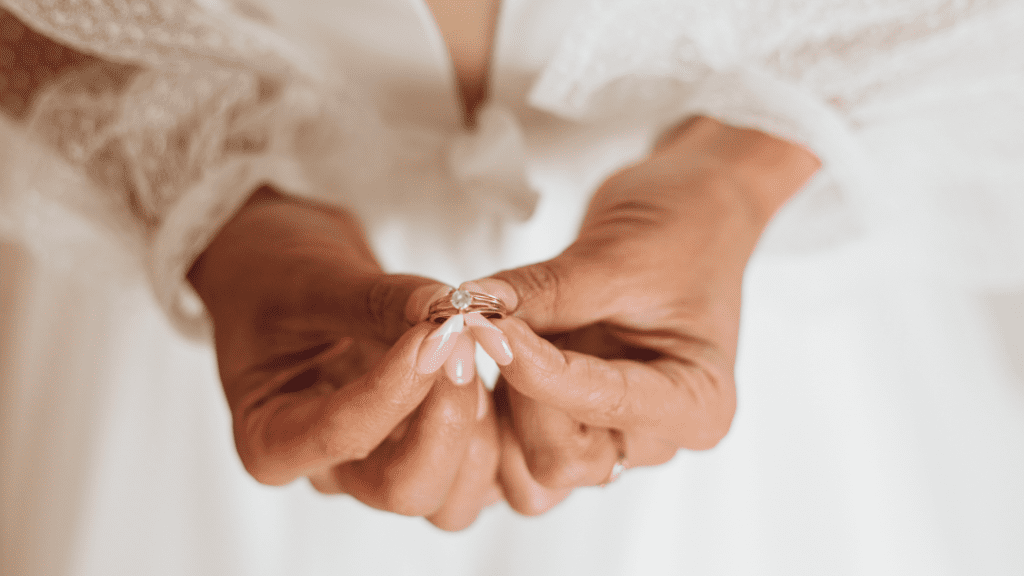 Bride with delicate pink nails holds a gold engagement ring with a sparkling diamond, dressed in a lace gown, preparing for her wedding ceremony.