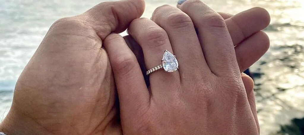 A romantic close-up of a couple holding hands, showcasing a pear-shaped diamond engagement ring with a glittering band against a serene waterfront backdrop.