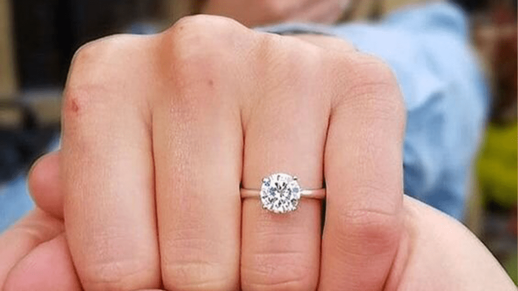 Close-up of a hand showing off a sparkling, round-cut diamond ring on the ring finger, with the person wearing a blue shirt in the blurred background.