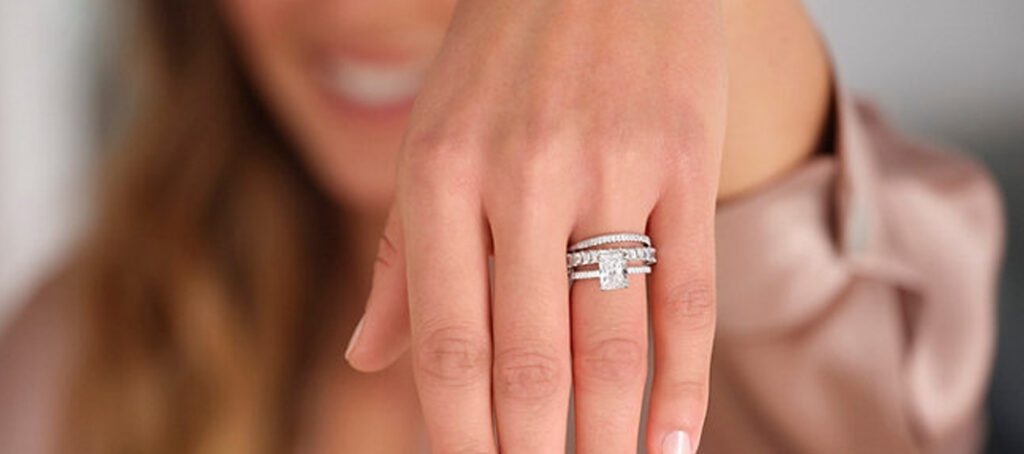 A woman showing off her hand adorned with a set of three stacked diamond rings, including a princess-cut engagement ring, smiling in the background.