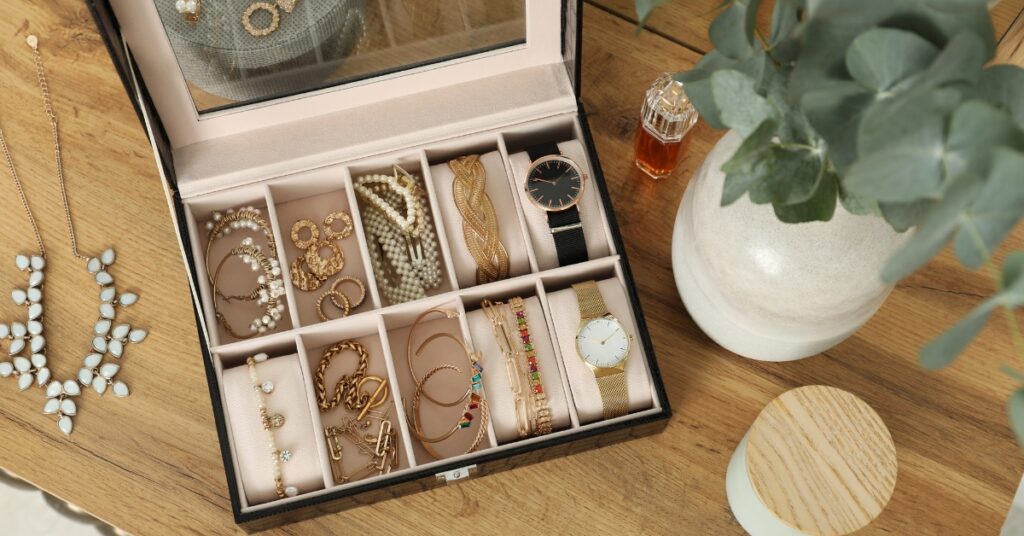 An organized jewelry box with various gold and pearl pieces, including bracelets, watches, and necklaces, next to a vase and perfume on a wooden table.