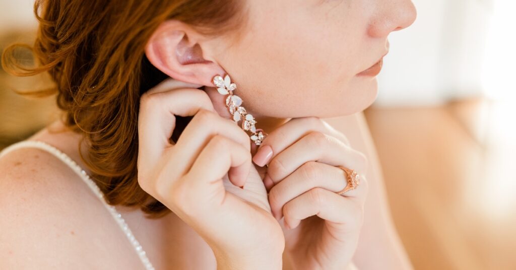 A woman with red hair delicately putting on a sparkling floral earring, showcasing her elegant jewelry and a rose gold ring on her finger.